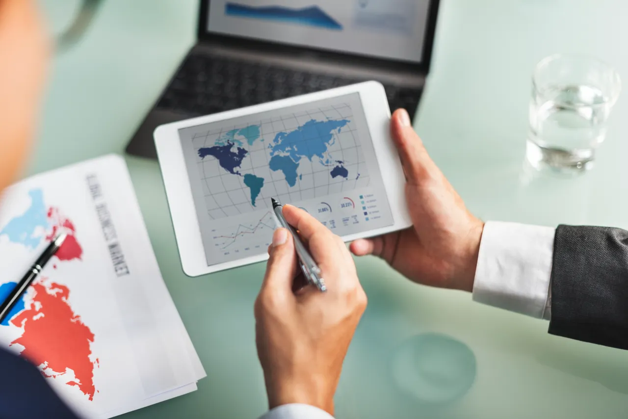 Mani di figure professionali al lavoro con una penna digitale su un tablet che mostra una mappa globale e grafici dei mercati esteri, a rappresentare i progetti di internazionalizzazione e marketing digitale del Bando Digital Export 2026-2027 dell'Emilia-Romagna.